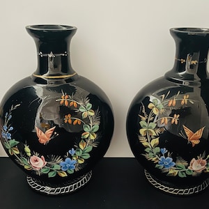 May include: Two black ceramic vases with gold trim and floral designs. The vases have a round body and a narrow neck. The floral designs include butterflies, roses, and leaves in shades of blue, pink, orange, and green.