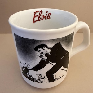 Puede incluir: Una taza de cerámica blanca con un estampado gráfico en blanco y negro de un artista sosteniendo un micrófono en la parte frontal. El borde interior de la taza muestra el nombre Elvis impreso en rojo. La taza tiene un asa curva en el lado derecho.