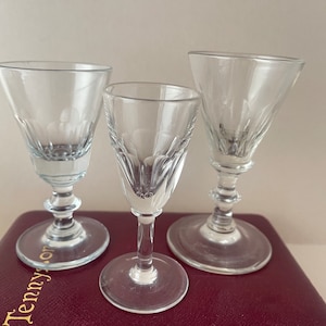 Puede incluir: Tres vasos de chupito de vidrio transparente con un patrón grabado decorativo. Los vasos tienen una base con tallo y están dispuestos sobre una superficie roja.
