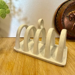 May include: A white ceramic toast rack with six arched slots and a handle. The rack sits on a wooden surface, with a copper wall hanging featuring a bull in the background. A green vase with greenery is on the left.