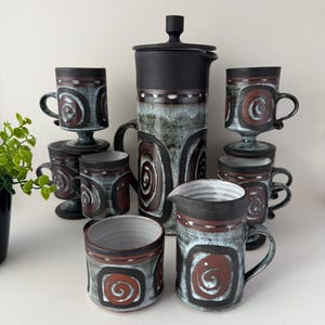 Puede incluir: Un juego de café de cerámica con una cafetera negra, una jarra para la crema y varias tazas. El juego tiene un diseño único con patrones de espirales marrones sobre un fondo azul claro y gris. Las tazas son de varios tamaños.