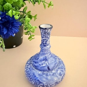 May include: A blue and white ceramic vase with a bulbous base and a narrow neck. The vase features a detailed landscape scene and floral patterns. The vase is displayed near a black pot with white polka dots and green and blue flowers.