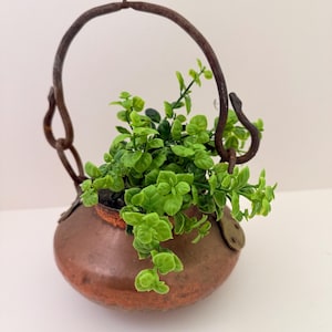 May include: A small, copper-coloured cauldron with a rusty metal handle and a decorative metal loop for hanging. The cauldron is filled with vibrant green artificial plants, creating a rustic decorative piece.