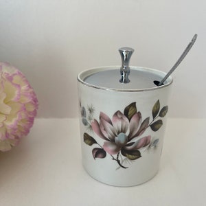 Peut inclure: Sucrier en céramique blanche avec un motif floral composé de fleurs roses et brunes. Le bol a un couvercle argenté avec un bouton et une cuillère argentée assortie.