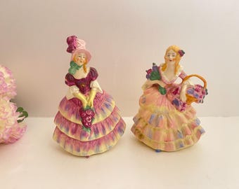 Peut inclure: Deux figurines en porcelaine vintage de femmes en robes élaborées. L'une porte un chapeau rose et tient des raisins, l'autre porte un panier de fleurs. Les robes sont rose pastel, jaune et violet.