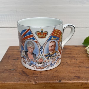 May include: White ceramic mug with a royal blue, red, and gold design featuring portraits of King George V and Queen Mary. The mug has a gold trim and the text "Dieu et mon Droit" on the bottom.