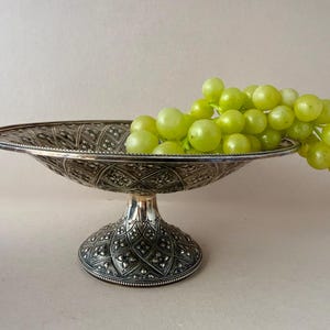 May include: A silver-toned metal pedestal bowl featuring an intricate embossed diamond pattern with small floral accents. A large cluster of fresh green grapes rests inside the bowl, with several grapes hanging over the edge of the rim.