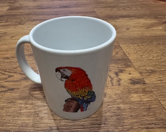 May include: White ceramic mug featuring a colorful illustration of a macaw parrot. The parrot has red, yellow, blue, and green feathers. The mug has a curved handle and is set against a wooden surface.