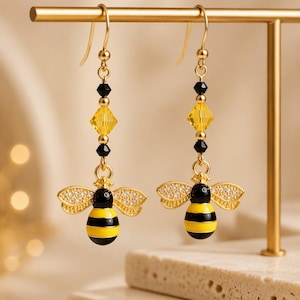 May include: A pair of gold-tone drop earrings featuring a bee charm with black and yellow enamel stripes and clear crystal wings. Above the bee, each earring has a faceted yellow crystal bead flanked by two small black beads on a gold wire hook.