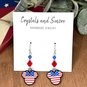 May include: Red, white, and blue American flag themed dangle earrings with crystal accents. The earrings feature a Mickey Mouse silhouette with a bow. The earrings are handmade and are labeled "Crystals and Smore Handmade Jewelry".