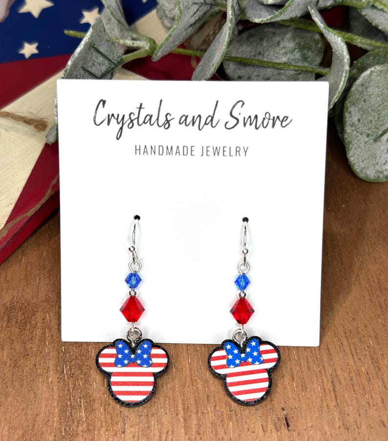 May include: Red, white, and blue American flag themed dangle earrings with crystal accents. The earrings feature a Mickey Mouse silhouette with a bow. The earrings are handmade and are labeled "Crystals and Smore Handmade Jewelry".