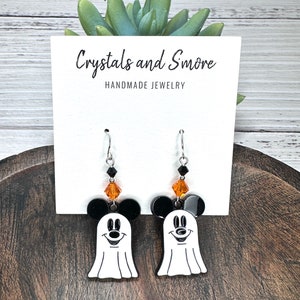 Puede incluir: Un par de pendientes de tem&aacute;tica de Halloween con un fantasma blanco con orejas de Mickey Mouse negras y detalles de cristal naranja y negro. Los pendientes est&aacute;n en una tarjeta blanca con el texto "Crystals and Smore Handmade Jewelry".
