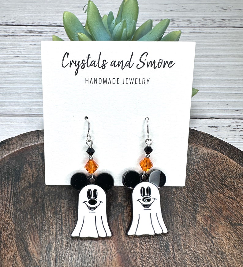 Puede incluir: Un par de pendientes de tem&aacute;tica de Halloween con un fantasma blanco con orejas de Mickey Mouse negras y detalles de cristal naranja y negro. Los pendientes est&aacute;n en una tarjeta blanca con el texto "Crystals and Smore Handmade Jewelry".