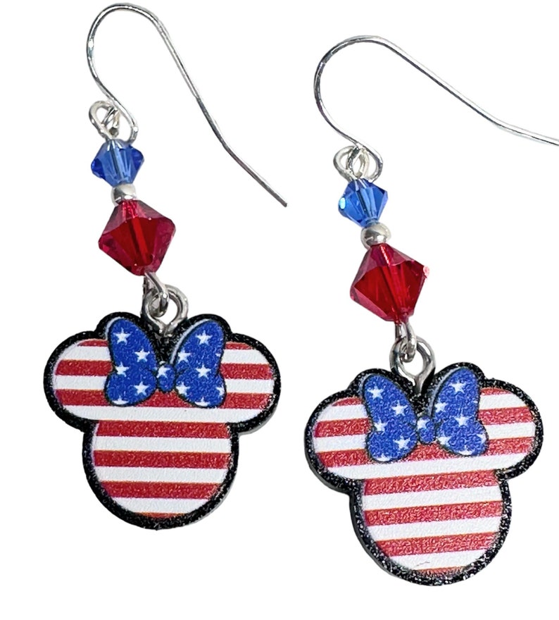 May include: A pair of silver dangle earrings featuring a red, white, and blue American flag design with a blue bow. The earrings are accented with red and blue crystal beads.