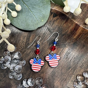 May include: A pair of silver dangle earrings featuring red, white, and blue American flag-themed Mickey Mouse ears with a red bow. The earrings have red and blue crystal beads.