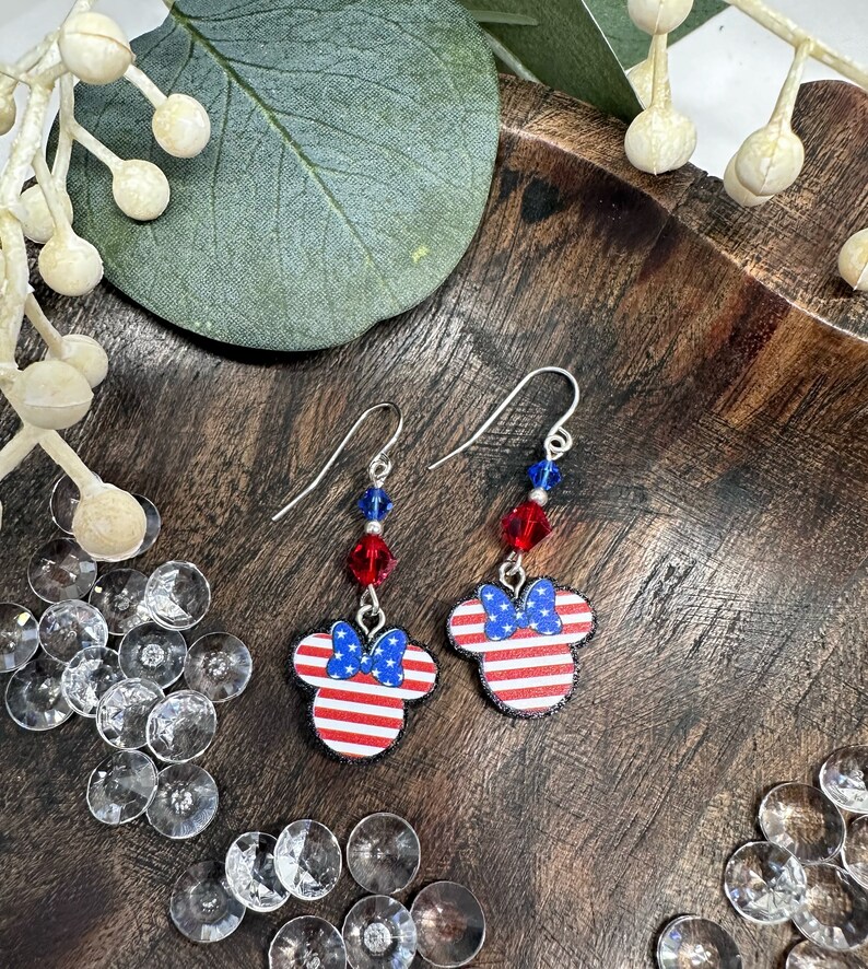 May include: A pair of silver dangle earrings featuring red, white, and blue American flag-themed Mickey Mouse ears with a red bow. The earrings have red and blue crystal beads.