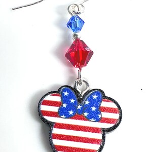 May include: Red, white, and blue American flag themed dangle earrings with a Mickey Mouse head silhouette and a blue bow. The earrings feature a red crystal and a blue crystal.
