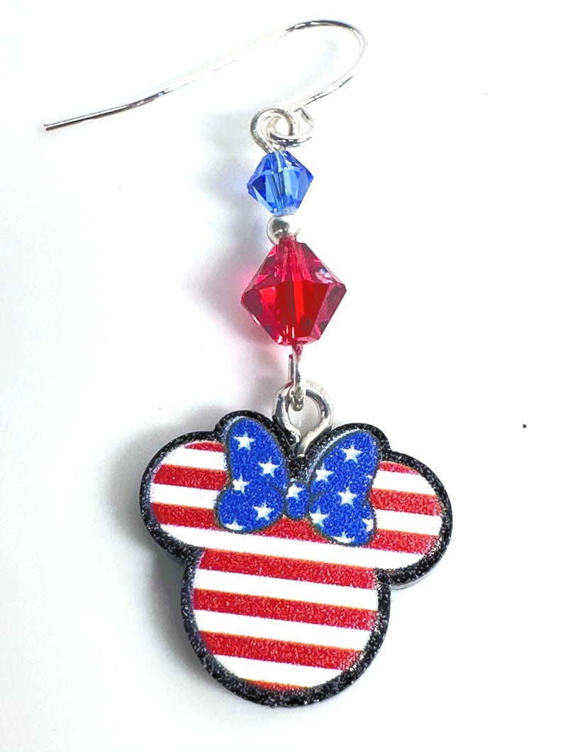 May include: Red, white, and blue American flag themed dangle earrings with a Mickey Mouse head silhouette and a blue bow. The earrings feature a red crystal and a blue crystal.