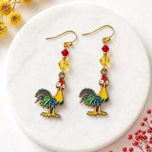 May include: A pair of dangle earrings featuring enameled rooster charms with yellow, blue, and green details. Each earring includes a faceted red crystal bead and a faceted yellow crystal bead suspended from a gold-toned metal hook wire.