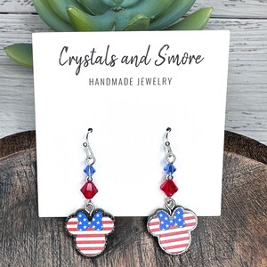 May include: Red, white, and blue American flag themed dangle earrings with a silver hook and crystal accents. The earrings feature a Mickey Mouse silhouette with a bow. The card reads "Crystals and Smore Handmade Jewelry".