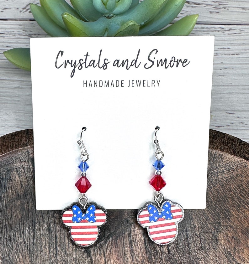 May include: Red, white, and blue American flag themed dangle earrings with a silver hook and crystal accents. The earrings feature a Mickey Mouse silhouette with a bow. The card reads "Crystals and Smore Handmade Jewelry".