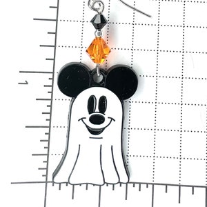 Puede incluir: Fantasma blanco y negro con orejas de Mickey Mouse y una cara sonriente. El fantasma lleva una cuenta negra y una cuenta de cristal naranja.