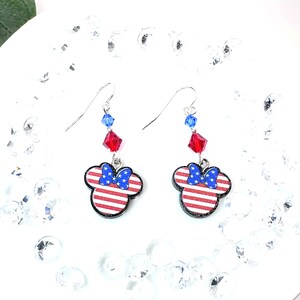 May include: A pair of dangle earrings featuring a red, white, and blue American flag design with a blue bow on a Mickey Mouse head silhouette. The earrings are accented with red and blue crystal beads.