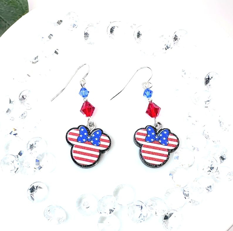 May include: A pair of dangle earrings featuring a red, white, and blue American flag design with a blue bow on a Mickey Mouse head silhouette. The earrings are accented with red and blue crystal beads.