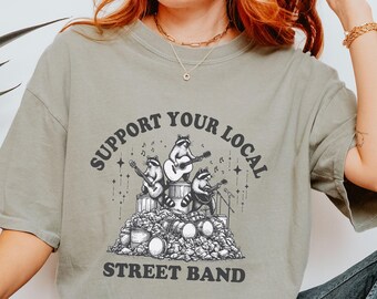 May include: A light green t-shirt featuring a graphic of raccoons playing music on a pile of rubbish. The text "Support Your Local Street Band" is printed on the shirt.