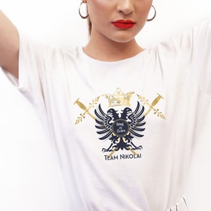 Puede incluir: Camiseta blanca con un escudo dorado y negro con dos águilas, una corona y espadas. El texto "Sisters in Law" y "TEAM NIKOLAI" están en el escudo.