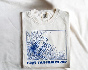 May include: White t-shirt with a blue illustration of a frog in a pond. The text "rage consumes me" is printed below the image.