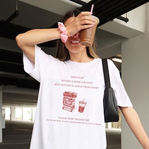 May include: White t-shirt with the text "THIS IS MY BUYING A BOOK (OR FIVE) AND GETTING A LITTLE TREAT SHIRT" in red. Below the text is a graphic of a stack of books and a drink. The shirt also says "PLEASE DONT BOTHER ME I'M ON MY WAY TO BUY A BOOK AND GET A LITTLE TREAT."