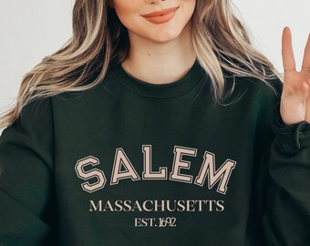 Könnte beinhalten: Ein dunkelgrüner Pullover mit dem Text "SALEM MASSACHUSETTS EST. 1692" in hellrosa.