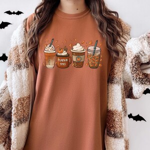 May include: A brown long-sleeved shirt with a graphic of four fall-themed coffee drinks. The drinks are labeled "Pumpkin Spice" and have fall-colored leaves and pumpkins around them.