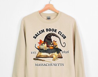 Könnte beinhalten: Ein beiger Pullover mit einem schwarzen Grafikdruck eines Hexenhutes, eines Buches und Kerzen. Der Text "Salem Book Club Est. 1626 Massachusetts" ist unter dem Grafikdruck gedruckt.