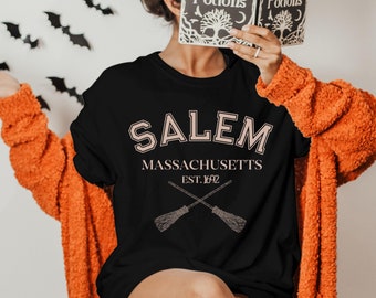 May include: Black long-sleeved t-shirt with a pink graphic print that says "SALEM MASSACHUSETTS EST. 1692" with two crossed broomsticks underneath the text.