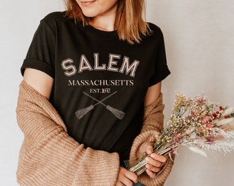 Puede incluir: Una camiseta negra con el texto "SALEM MASSACHUSETTS EST. 1692" y un gráfico de dos escobas cruzadas.