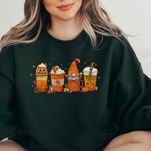 May include: A dark green sweatshirt featuring a graphic of four autumn-themed drinks. The drinks are decorated with pumpkins, leaves, and a gnome.