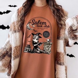 Puede incluir: Una sudadera marr&oacute;n con un dise&ntilde;o gr&aacute;fico de una bruja leyendo un libro frente a una pila de libros. El gr&aacute;fico tambi&eacute;n incluye una luna creciente, murci&eacute;lagos y el texto "Salem Book Club".