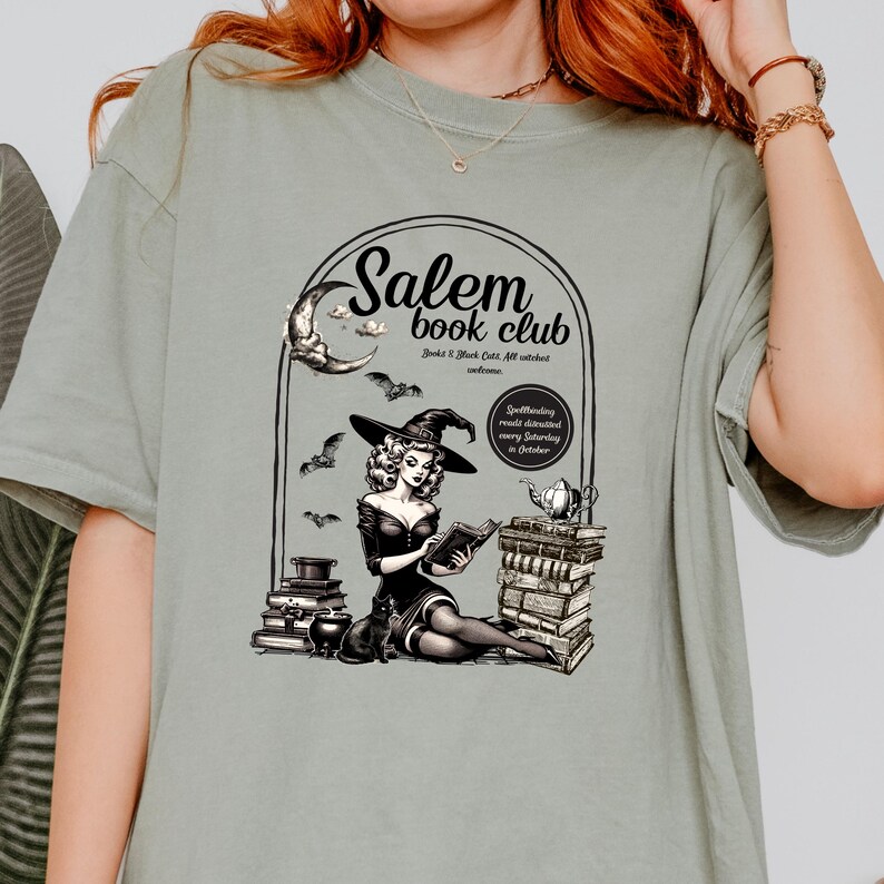 Puede incluir: Una camiseta verde con una ilustraci&oacute;n vintage de una bruja leyendo un libro frente a una pila de libros. El texto en la camiseta dice "Salem book club" y "Books & Black Cats. All witches welcome." El texto tambi&eacute;n dice "Spellbinding reads discussed every Saturday in October."