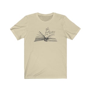 May include: A beige t-shirt with a black and white illustration of an open book. The text "the book was better" is written above the book.