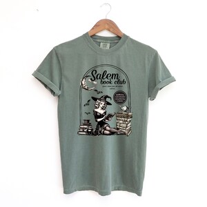 Puede incluir: Una camiseta verde con un gr&aacute;fico de una bruja leyendo un libro frente a una chimenea. El gr&aacute;fico tambi&eacute;n incluye el texto "Salem book club".