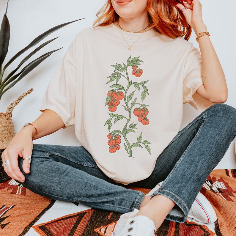 May include: A cream colored t-shirt with a graphic of a tomato plant with red tomatoes. The person wearing the shirt is sitting on a patterned rug.