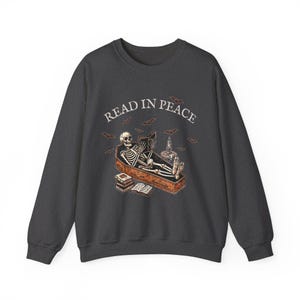 May include: A dark gray sweatshirt with a skeleton reading a book in a coffin. The text "READ IN PEACE" is above the skeleton.