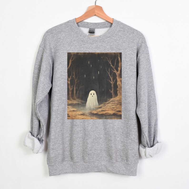 Puede incluir: Una sudadera gris con un estampado gr&aacute;fico de un fantasma blanco en un bosque oscuro. El fantasma est&aacute; de pie en un charco de agua con lluvia cayendo a su alrededor.