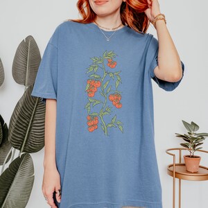 May include: A blue oversized t-shirt with a graphic of a tomato plant with red tomatoes. The plant has green leaves and stems.