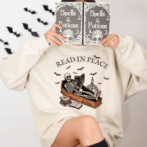 May include: A cream colored sweatshirt with a skeleton reading a book in a coffin. The text "Read in Peace" is above the skeleton. The sweatshirt has a crew neck and long sleeves.