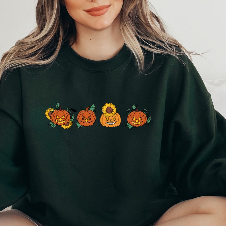May include: A dark green sweatshirt featuring a line of five orange jack-o'-lantern pumpkins with smiling faces. The pumpkins are adorned with sunflowers and leaves.