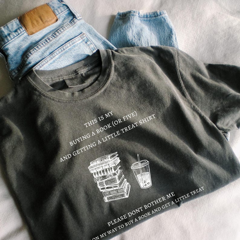 May include: Dark olive green t-shirt with white text and illustrations. The shirt reads "THIS IS MY BUYING A BOOK (OR FIVE) AND GETTING A LITTLE TREAT SHIRT." A stack of books and a drink are illustrated below the text. Light blue jeans are in the background.