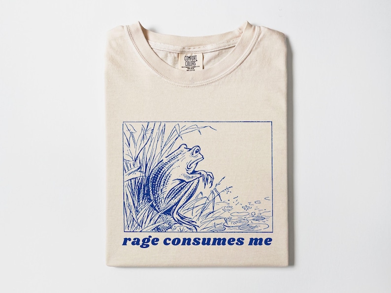 May include: A beige t-shirt with a blue illustration of a frog in a pond and the text "rage consumes me" printed on the front.